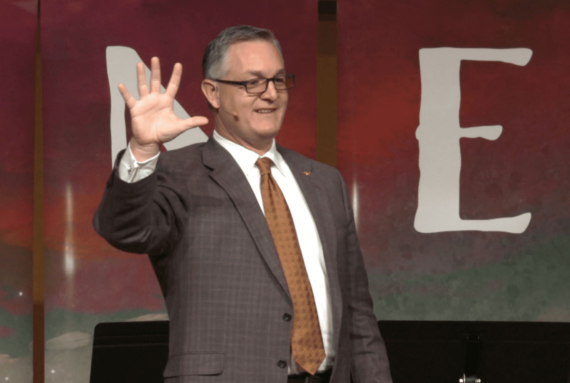 John Ramstead on stage, raising a hand to the audience